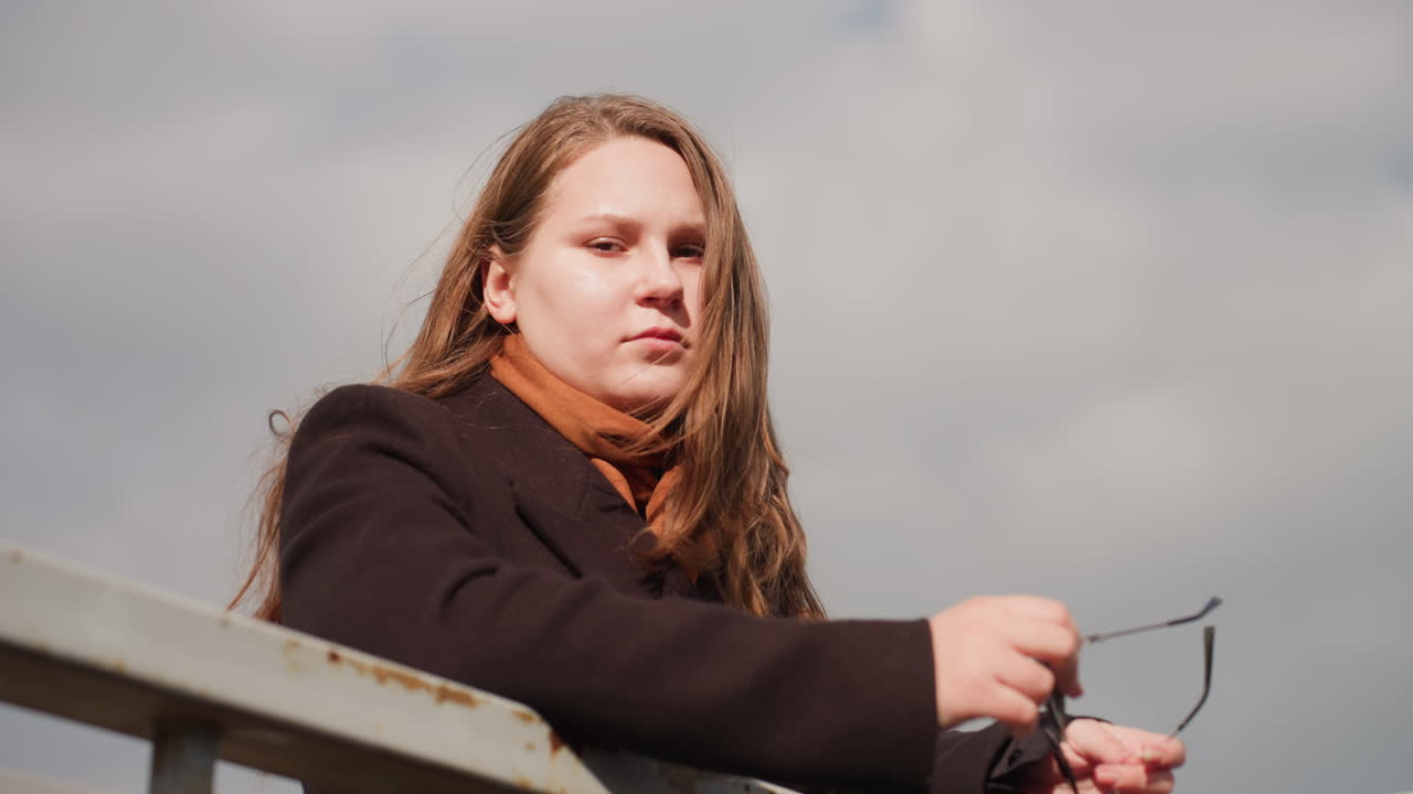 Soft morning sunlight glows gently on woman standing near metal railing, facing camera direction with calm expression, highlighting warmth, freshness, natural beauty, and peaceful outdoor emotion