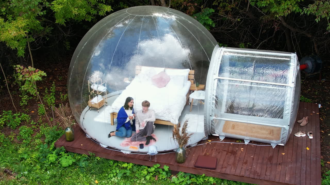 Transparent bubble tent at glamping, Lush forest around and interior. A couple sitting under a blanket, holding cups and talking
