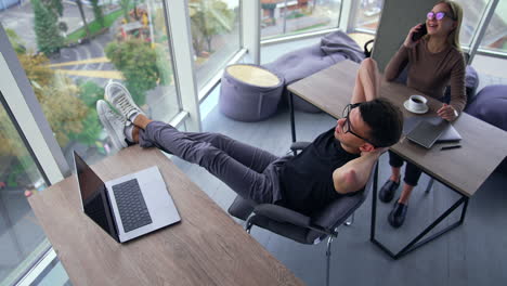 people having a break from work in the office. man sits with his legs on the table and hands behind his head. woman next table speaking on the phone. high angle view. city in window.