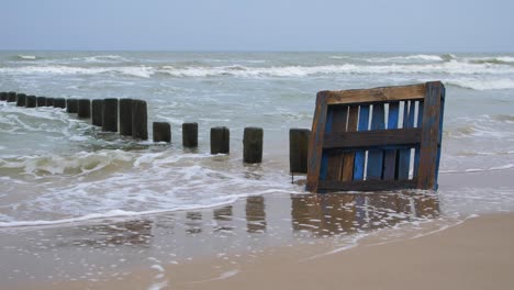 Old-blue-wooden-EPAL-palette-on-the-shore,-trash-and-waste-litter-on-an-empty-Baltic-sea-white-sand-beach,-old-wooden-pier,-environmental-pollution-problem,-overcast-day,-medium-shot