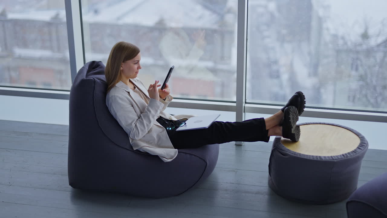 Calm relaxed lady looks at her phone sitting at chair stretching legs in front of her. Woman has laptop and glasses on her laps. Snow falling behind the window at backdrop.
