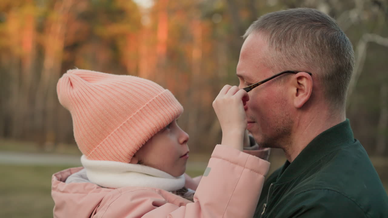 초록색 재을 입은 남자가 분홍색 재과 베니를 입은 어린 소녀를 안고있는 가 따뜻한 순간. 아이는 부드러운 상호 작용을 공유하면서 남자의 안경을 장난스럽게 조정하고 있습니다.