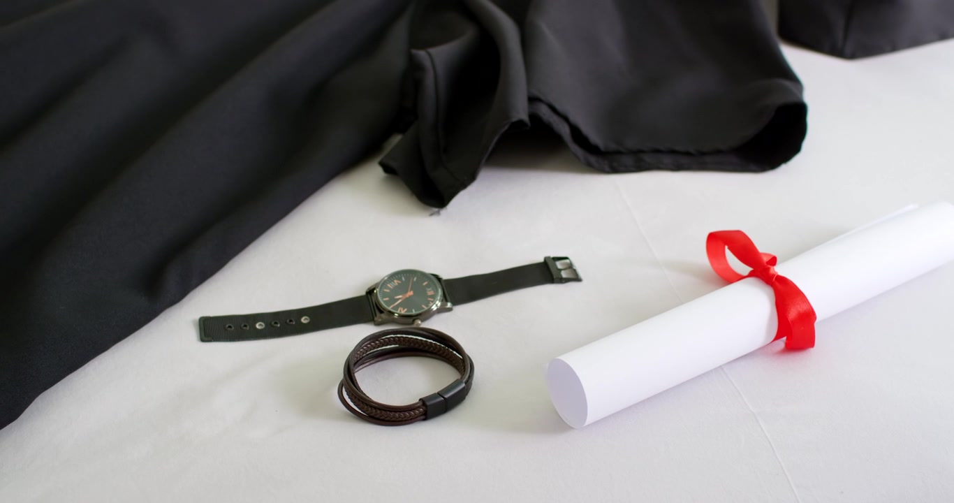 Telecamera che esegue panoramica e zoom rivelando abito nero, orologio, braccialetto e diploma su tessuto bianco per la preparazione alla laurea.