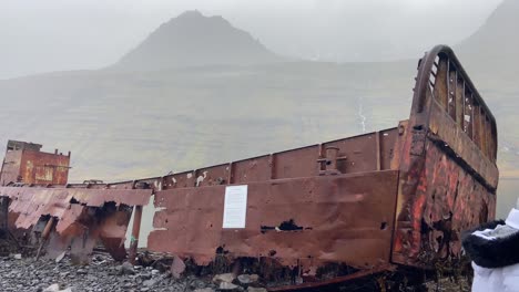 Happy-blonde-tourist-walking-by-rusty-old-shipwreck-stranded-Mjoifjordur,-East-Iceland