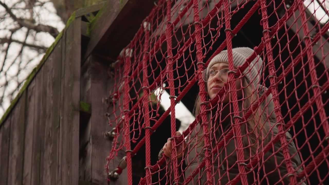 Elderly white woman behind red netting, grandmother peering from weathered treehouse, knitted beanie and coat, hands gripping rope mesh, cold overcast sky, mossy wooden beams, nostalgic introspective