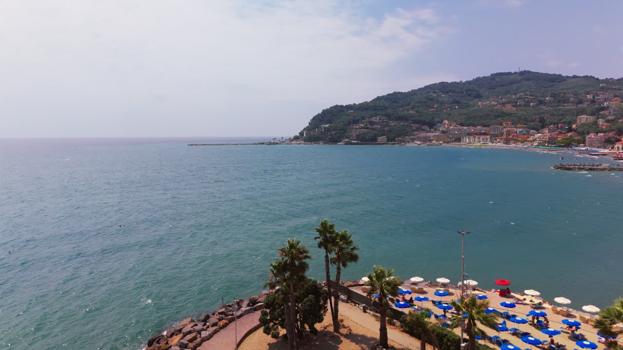 Low drone shot flying over a small palm-lined promontory and umbrellas on the beach, then advancing over the sea in Varigotti, Liguria