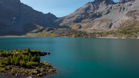 Aerial-flyover-alongside-an-island-in-the-waters-of-Lac-de-Salanfe-in-Valais,-Switzerland-on-a-sunny-autumn-day-in-the-Swiss-Alps-with-a-pan-down-view-from-the-surrounding-alpine-peaks-and-cliffs