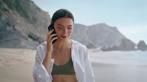 Girl-tourist-talking-mobile-walking-on-beach-near-stony-hills-close-up-vertical.