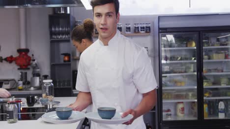 Mixed-race-male-cook-working-in-a-restaurant-kitchen,-holding-plates-with-food,smiling-at-the-camera