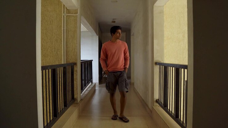 Young Indian man walking thoughtfully through an apartment corridor with hands in pockets, conveying solitude, contemplation, and everyday lifestyle.