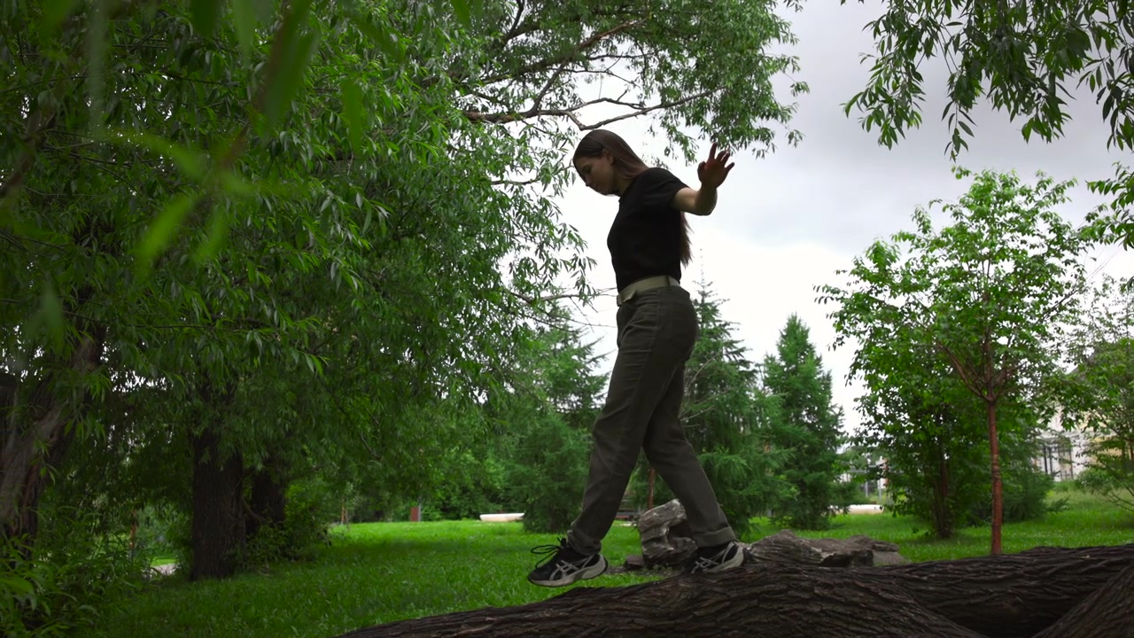 Full-body view of youth balancing and walking on large fallen tree trunk in landscaped urban park, surrounded by green foliage, grass, soft diffuse light, and calm outdoor setting during quiet
