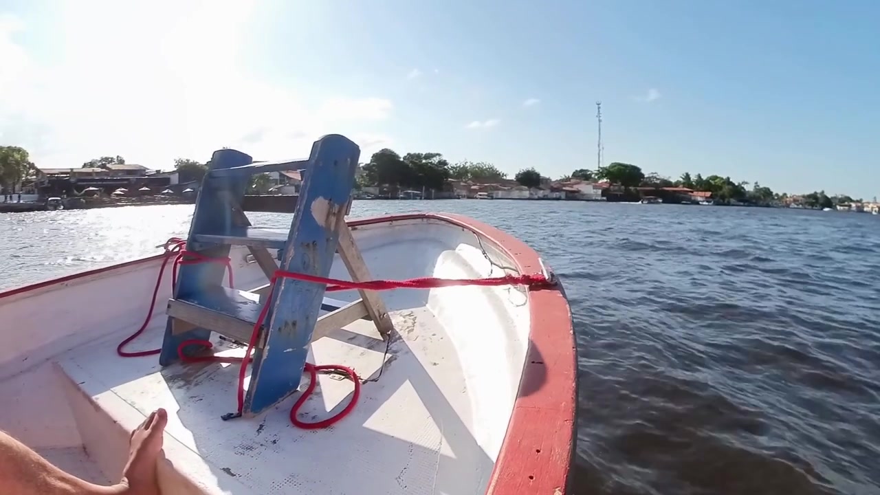 Inquadratura in soggettiva dalla prua di un motoscafo in legno che naviga le acque del fiume Preguiça nel Maranhão, Brasile, con edifici lungo il fiume e una vegetazione lussureggiante sotto la luce del sole