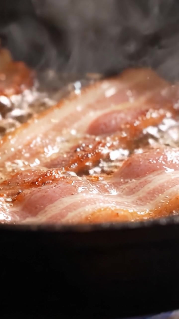 Pancetta sfrigolante che cuoce in padella: Un primo piano di strisce deliziosamente croccanti che friggono alla perfezione per una gustosa esperienza di pancetta.