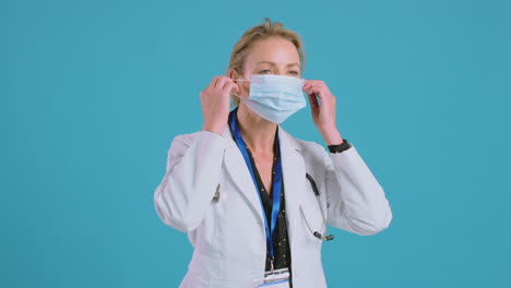 Portrait-Of-Female-Doctor-Wearing-White-Coat-Putting-On-Mask-In-Front-Of-Blue-Studio-Background