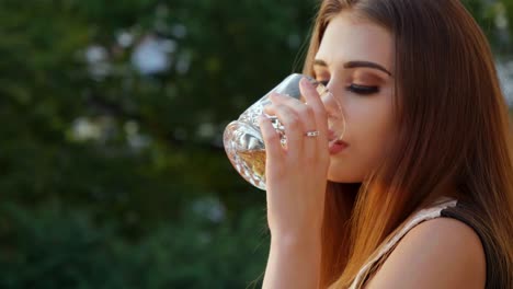 Plano-Medio-De-Una-Hermosa-Mujer-De-Moda-Mirando-A-La-Cámara-Mientras-Toma-Un-Trago-De-Whisky-De-Un-Vaso-De-Cristal-Mientras-Está-De-Pie-Afuera