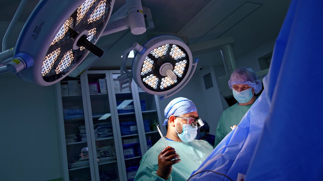 Male surgeons operate patient under the round lamps on the ceiling. Surgeons in surgical loupes uses metal tool and thread at operation.