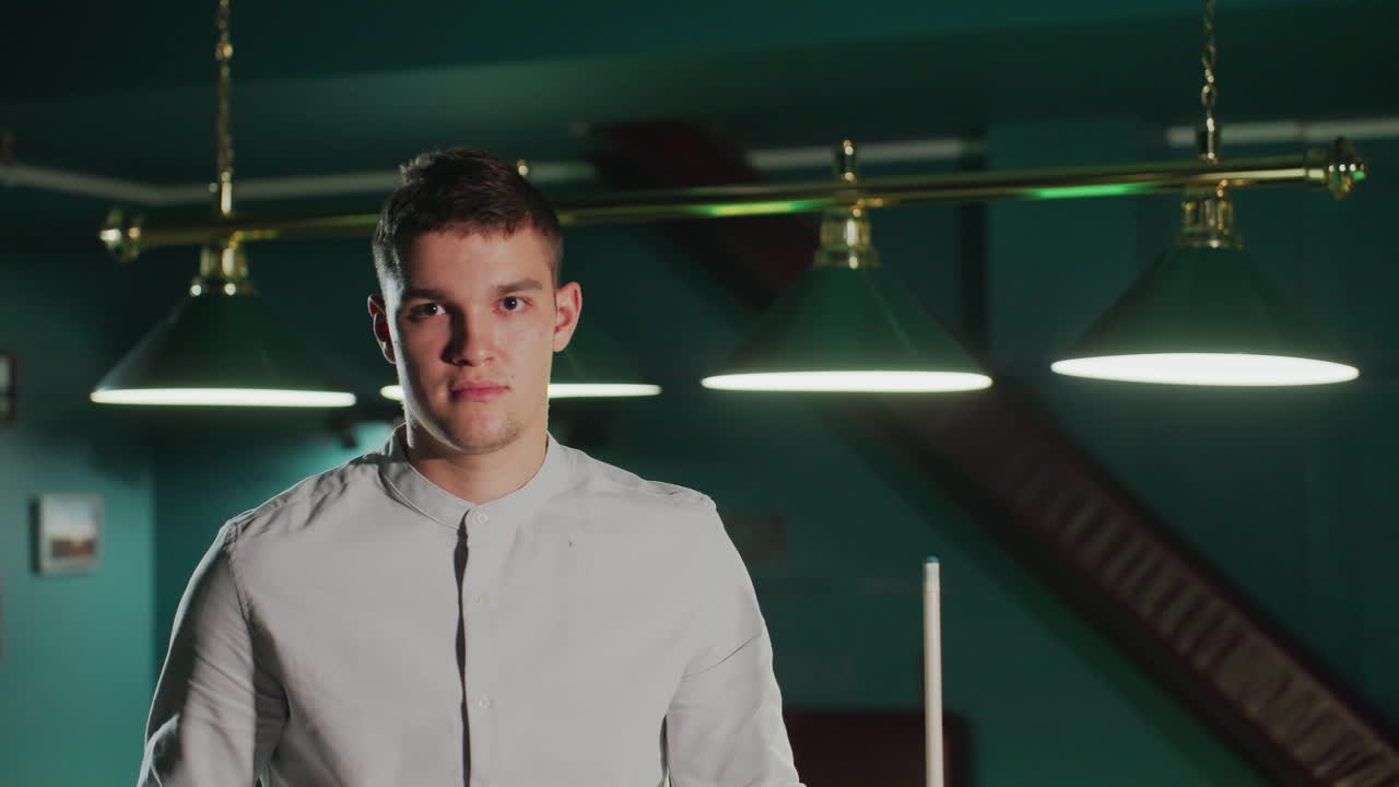 셔츠를 입은 남자가 진지한 표정으로 희미하게 조명된 수영장 홀에 서기 전에 큐 스틱을 들고 앉아 있습니다. 집중된 시선과 강렬한 분위기는 경쟁 정신과 집중력을 강조합니다.