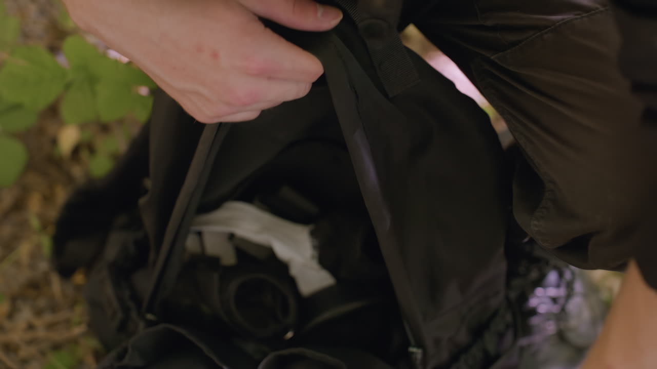 Adventurer pauses on leafy forest path, boots planted firmly on earthy ground, about to unzip black backpack containing visible equipment, surrounded by dry leaves, green plants and withered branches