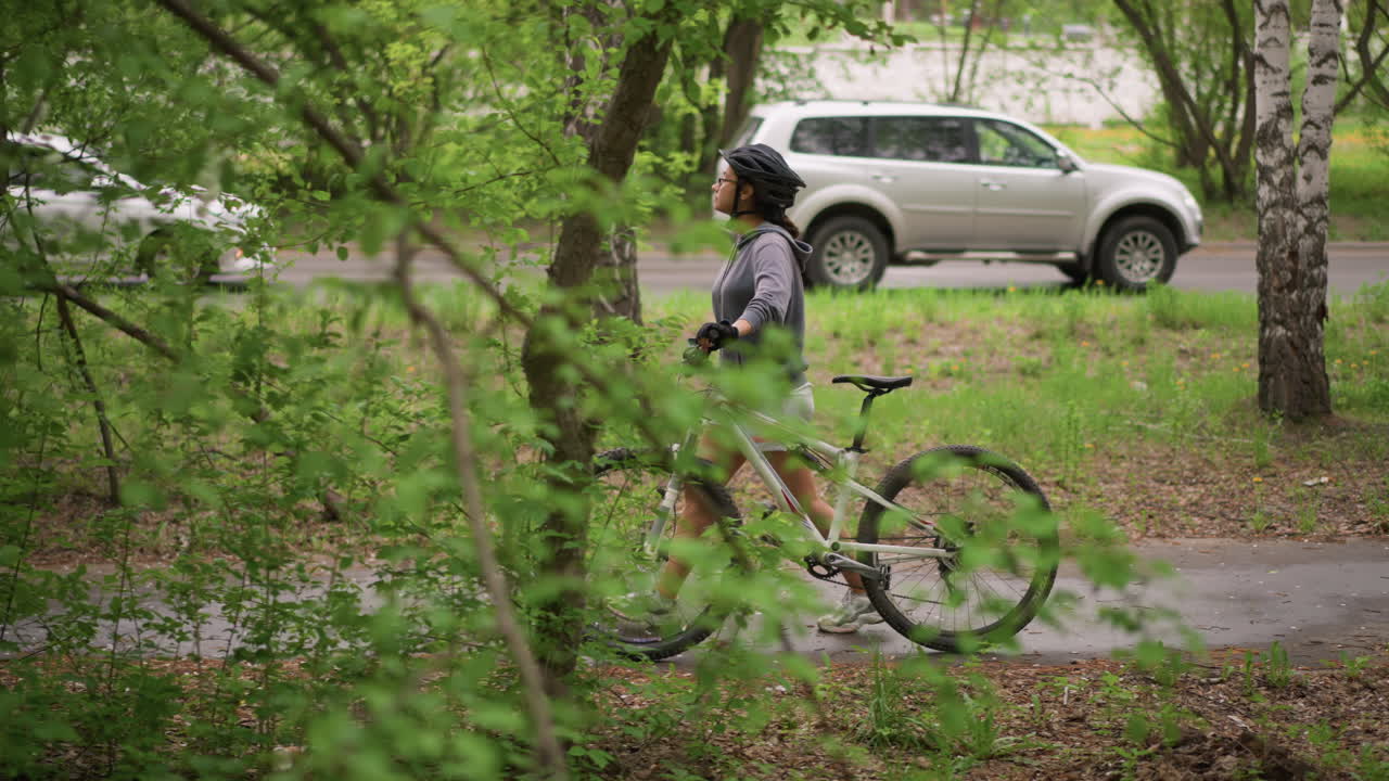 Woman Stops Bicycle Next To Road, Woman In Safety Gear Pauses With Her Bicycle Near Traffic And Greenery, Female Cyclist Taking Break Beside Her Bicycle With Passing Cars And Lush Trees Nearby