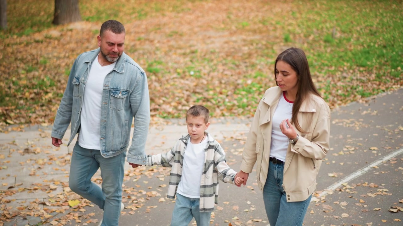 Famiglia di tre persone che cammina insieme tenendosi per mano in un parco con foglie autunnali a terra. Uomo, donna e bambino si muovono lungo un sentiero lastricato circondati da erba verde e foglie marroni cadute. Luce naturale soffusa.