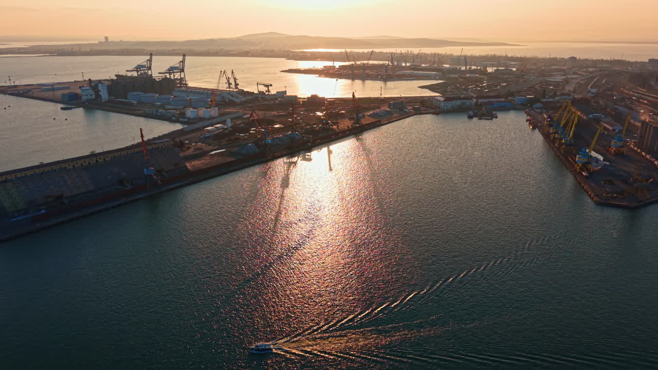 Sunset at the harbor with small boat moving through water near shipping docks and city skyline in the background