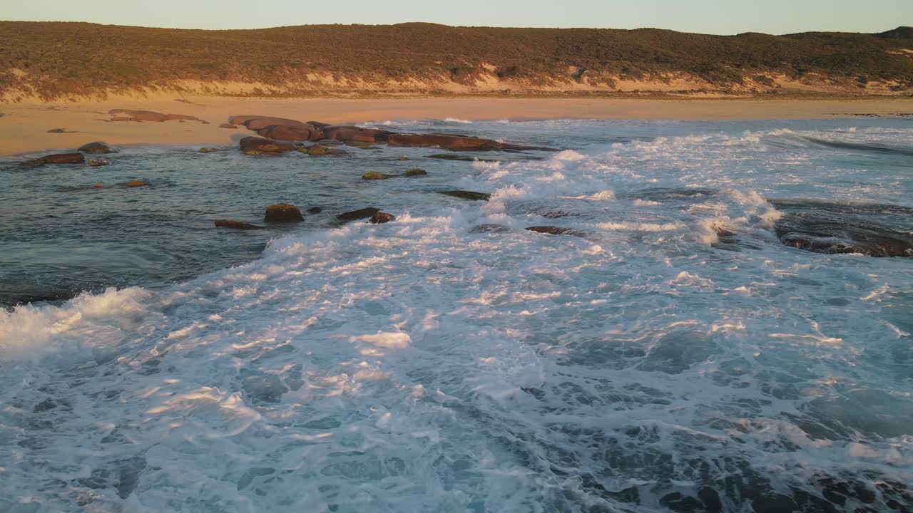 vuelo aéreo hacia atrás sobre salpicaduras de agua del océano chocando contra rocas y terminando en una playa de arena durante la puesta de sol dorada