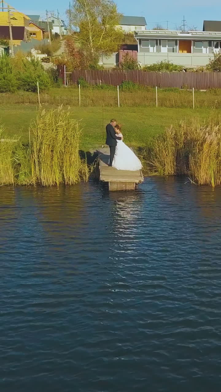 young newlywed couple hugs on small pier near large calm river against cottage town on sunny autumn day aerial view