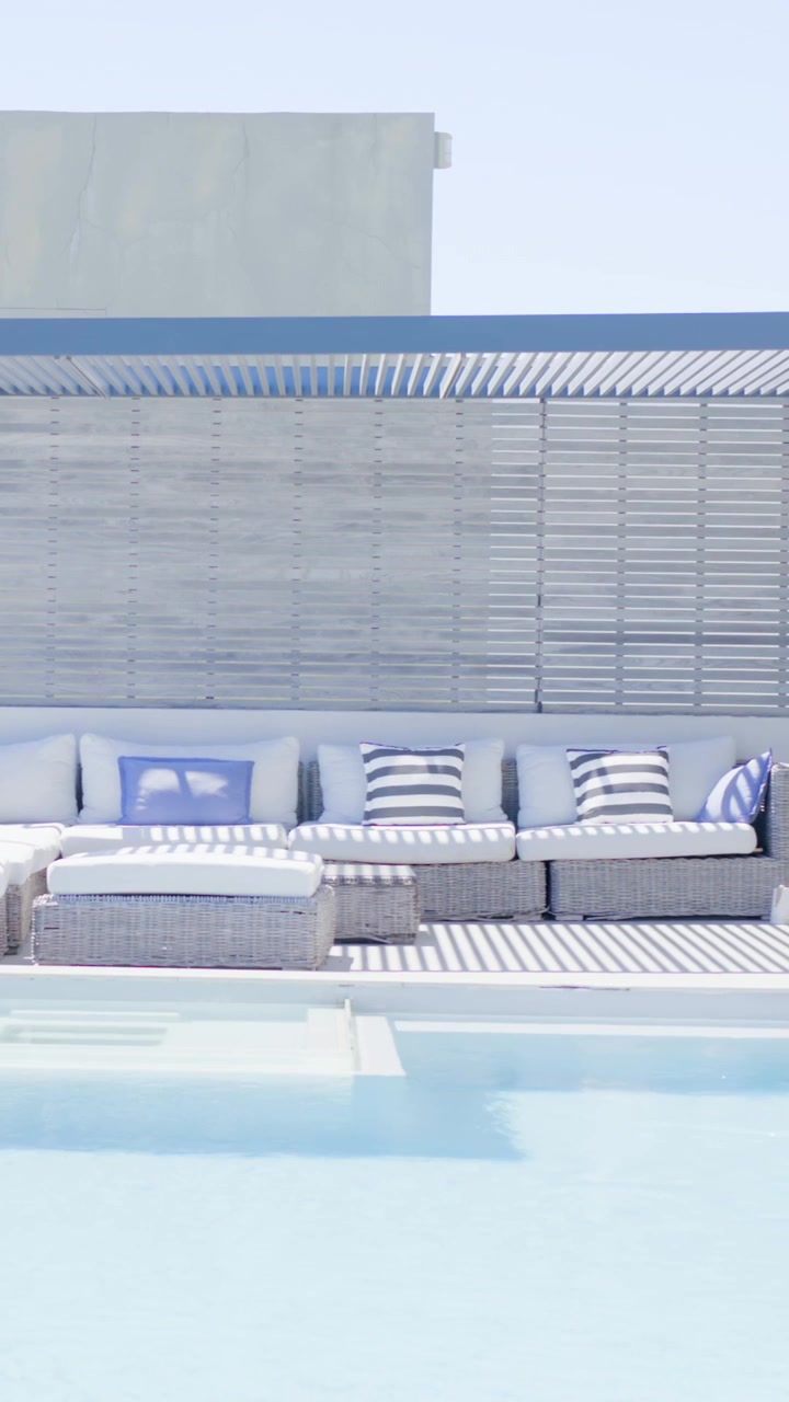 Vertical video: Shifting sunlight causing pool water rippling by wicker sofa with ottoman, pillows