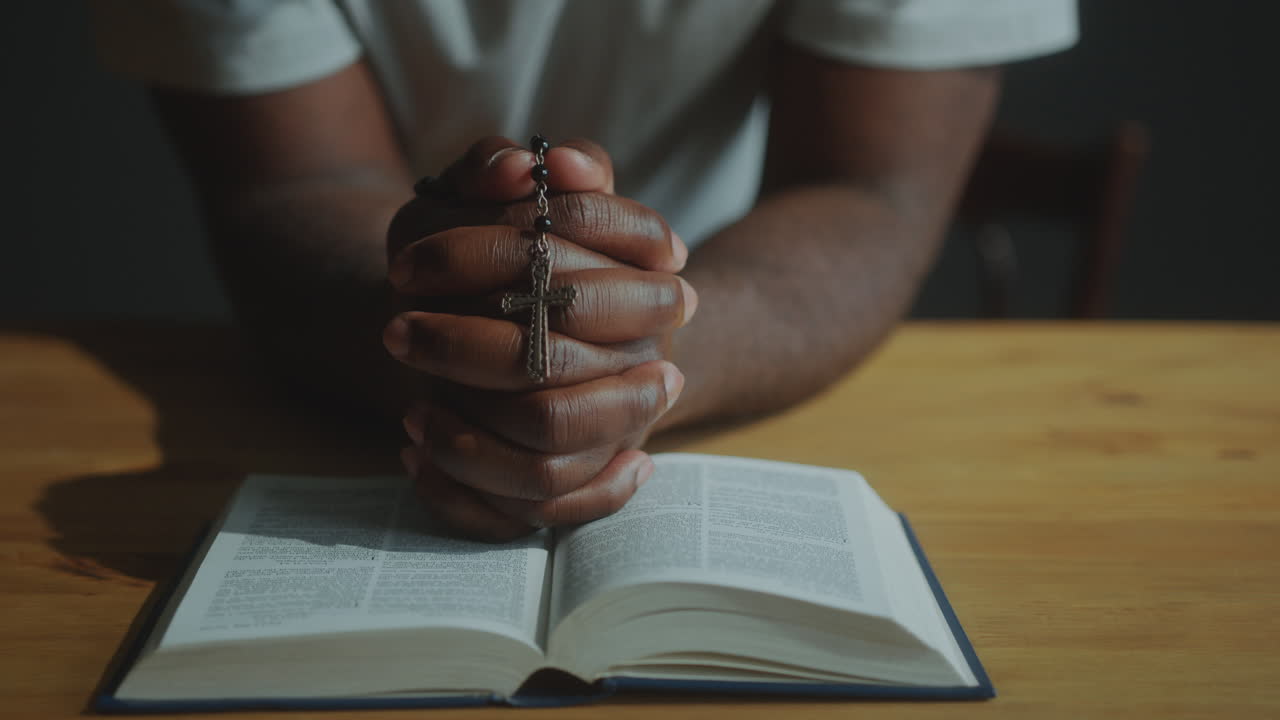 Hands of Black Man Holding Rosary Cross and Praying with Bible