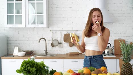 cheerful girl eats a banana in a white kitchen with a table with vegetables and fruits