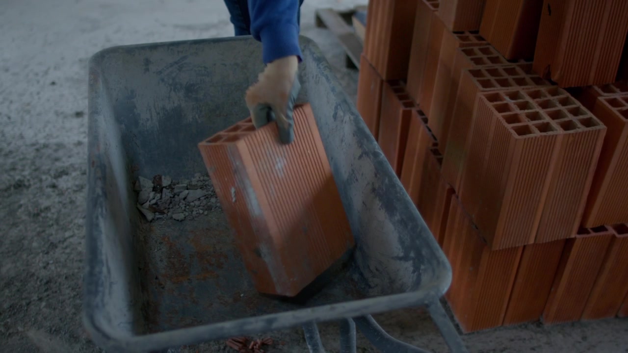 4K slow-motion footage of a construction worker in a safety vest and gloves removing red bricks from a wheelbarrow at a construction site, showcasing labor and precision.