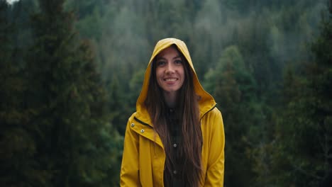 a happy girl in a yellow jacket stands and smiles against the backdrop of a green coniferous forest in the mountains during the rain