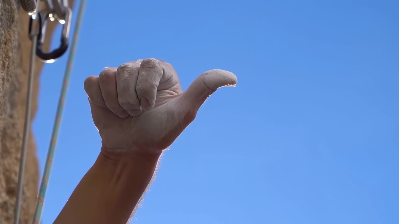 Un solo antebrazo y mano dan aprobación junto a equipo de escalada de piedra texturizada y metal, con un cielo azul saturado de fondo, luz solar nítida, sombras suaves y espacio en blanco limpio al aire libre