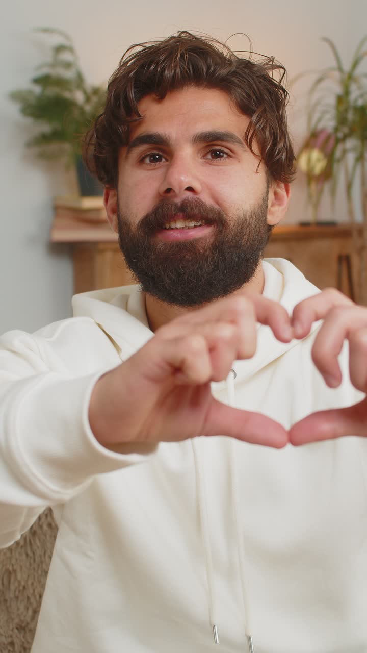 Happy indian young man at home living room couch makes symbol of love showing heart sign to camera