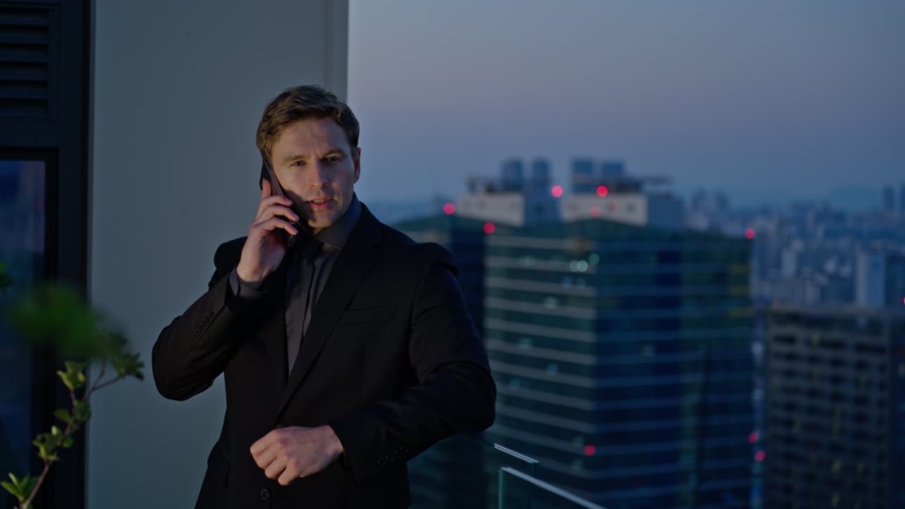 A Caucasian businessman in black suit having phone call at rooftop terrace with city skyline at sunset, smiling positive and confident man