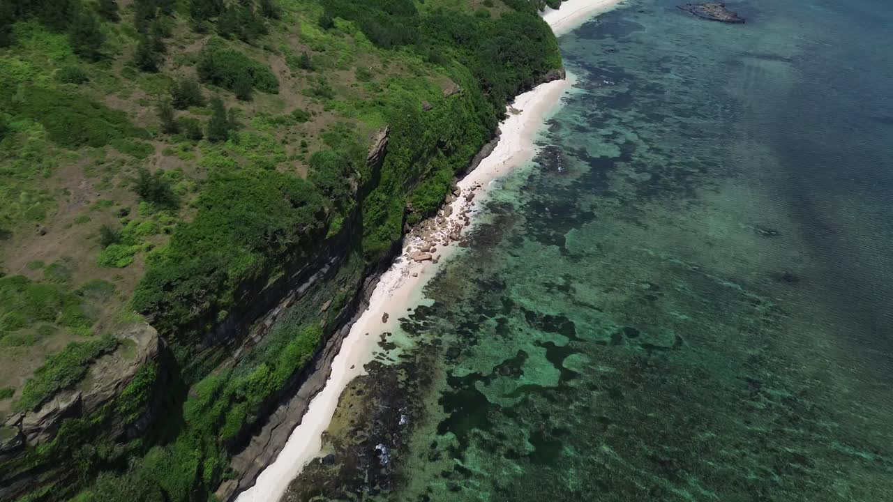 Zoom in following tropical coastline with clear water and steep green cliffs, captured on a sunny day in Ly Son Island