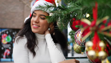 Una-Mujer-Joven-Con-Sombrero-De-Santa-En-La-Lista-Traviesa-Mientras-Cuelga-Adornos-Y-Cascabeles-En-El-árbol-De-Navidad