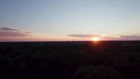 Drohnenaufnahmen-Zeigen-Den-Himmel-In-Einem-Orangefarbenen-Farbton,-Während-Die-Sonne-Am-Horizont-Untergeht-Und-Die-Kleine-Stadt-Von-Bäumen-Umgeben-Ist