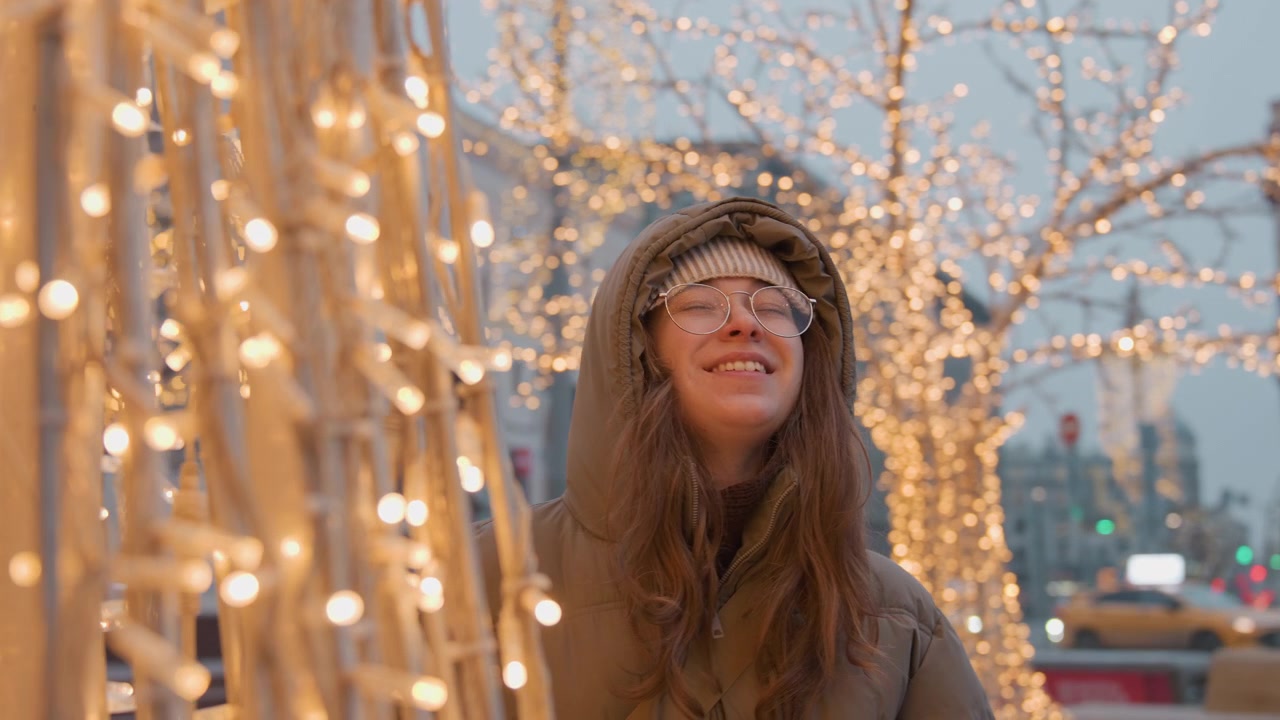 Ung kvinde i hættejakke kigger på gyldne lys på bytorv, blød bokeh-glød, kold skumring, historisk arkitektur, langsomt smil, filmisk stemning, hyggelig vinterstyling, fodgængerbaggrund