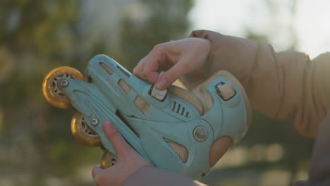 close-up shot of a light blue rollerblade being held and adjusted by a person in a sunlit park. the image captures the person's hands loosening the straps of the rollerblade, with a blurred background