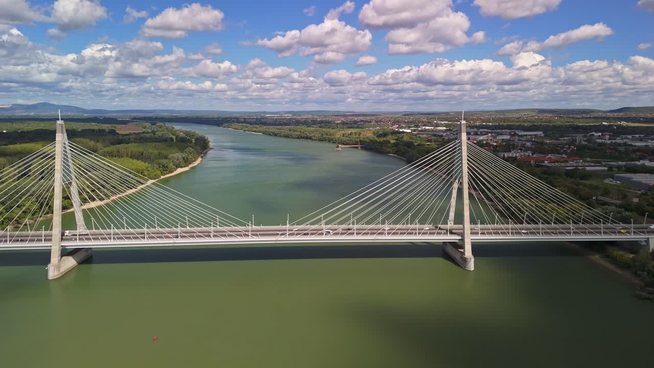 Aerial close-up of the Megyeri Bridge in Budapest, Hungary, showing its symmetrical cable-stayed design spanning the green waters of the Danube River under a bright, cloudy sky