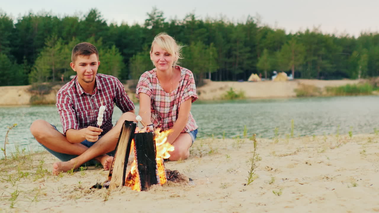 한 남자와 한 여자의 친구가 모닥불 주위에 앉아 막대기에 마시멜로를 굽고 있습니다.