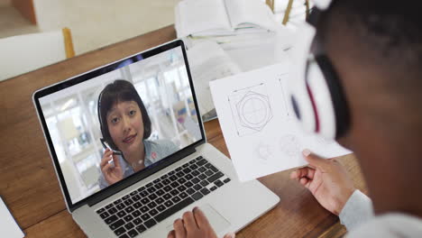 médico afroamericano que usa auriculares de teléfono tomando notas mientras hace una videollamada en una computadora portátil