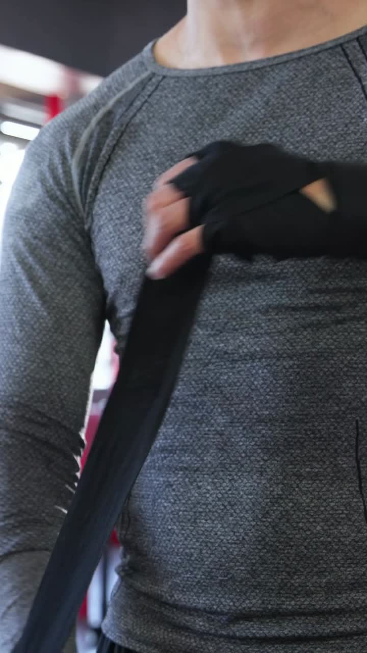 Vídeo vertical: Homem enrolando faixas de mão pretas na academia, preparando-se para treinar com uma blusa cinza.