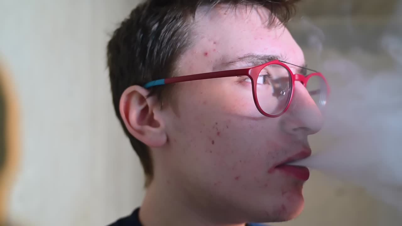 Young pimply man in red glasses vaping and exhales smoke indoor. Side view. Close up. Slow motion