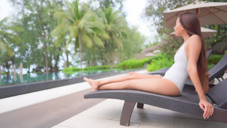 Mujer-Asiática-Esbelta-En-La-Silla-De-La-Piscina-Junto-A-La-Piscina-Del-Resort-Tropical-De-Lujo,-Fotograma-Completo