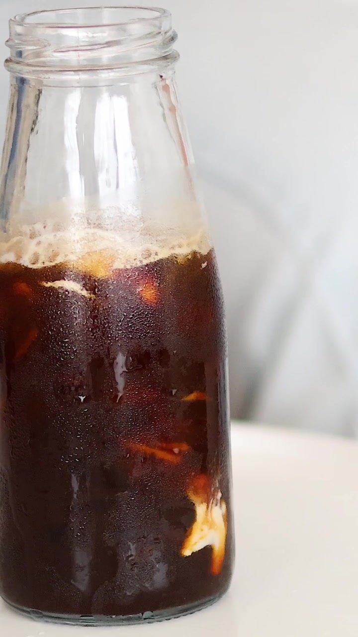 Una ripresa verticale che mostra panna che viene versata in una bottiglia di vetro piena di caffè scuro.