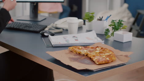 primer plano del gerente ejecutivo tomando una rebanada de pizza comiendo frente a la computadora escribiendo gráficos financieros