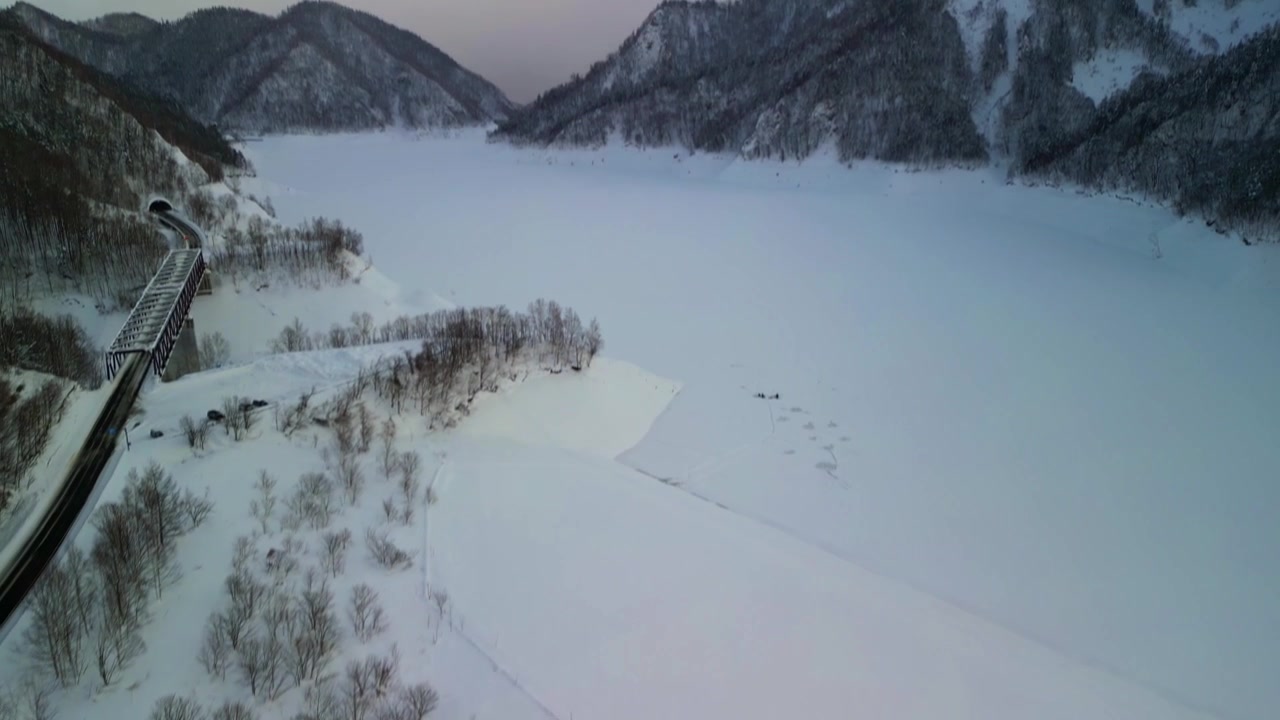 Droneflyvning over en tett snødekt skog som åpner seg og avslører en massiv frossen hvit innsjø og en jernbanebro om vinteren.