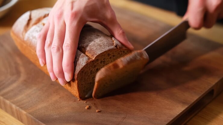 Hands Slicing Artisan Loaf On Board, Closeup Of Hands Using Serrated Knife To Cut Crusty Whole Grain Bread